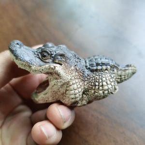May include: A detailed figurine of an alligator with an open mouth. The alligator is primarily gray and tan, with textured skin and a detailed head. The figurine appears to be made of ceramic or a similar material.