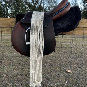 Puede incluir: Una silla de montar inglesa de cuero marrón con una almohadilla de piel de oveja negra y una cincha beige tejida. La silla de montar está colgada de una valla de alambre. Los estribos son plateados.