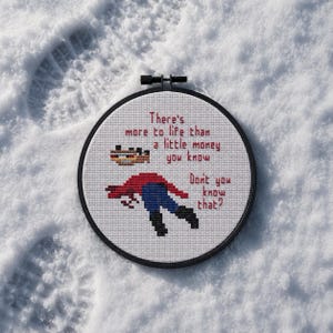May include: A circular black embroidery hoop containing white Aida fabric with a cross-stitch design. The text reads: There's more to life than a little money you know. Don't you know that? It features a pixelated figure in red and blue.