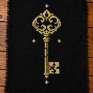 Könnte beinhalten: Eine Kreuzstichstickerei auf schwarzem Stoff mit einem zentralen, verzierten Schlüsselmotiv in Gold- und Gelbtönen. Der Schlüssel hat einen kunstvollen Kopf und einen rechteckigen Bart. Kleine Kreuzstich-Sterne befinden sich über und unter dem Schlüssel.