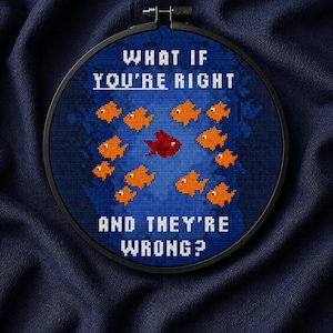 May include: A circular embroidery hoop containing a cross-stitch design on dark blue fabric. The design features eleven small orange fish swimming in one direction and one red fish swimming in the opposite direction. The text reads: WHAT IF YOU'RE RIGHT AND THEY'RE WRONG?