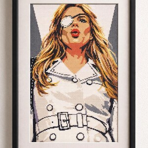Puede incluir: Obra de arte en punto de cruz enmarcada que presenta a una mujer con cabello rubio largo, un parche en el ojo y lápiz labial rojo. Lleva un abrigo blanco con cinturón y marco negro. Es un retrato.