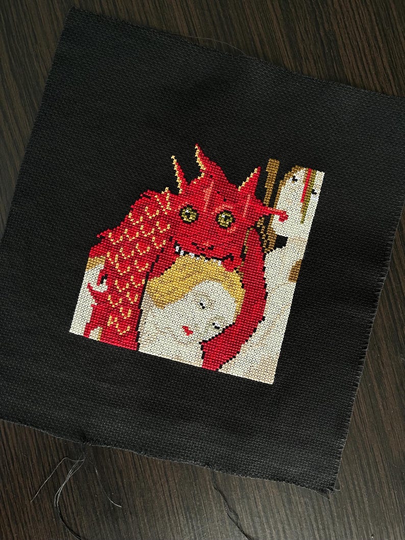 Pode incluir: Um peda&ccedil;o quadrado de tecido preto com um bordado em ponto cruz. A imagem retrata uma criatura vermelha e escamosa semelhante a um drag&atilde;o com olhos amarelos sobre uma figura de pele p&aacute;lida com cabelo loiro e l&aacute;bios vermelhos.