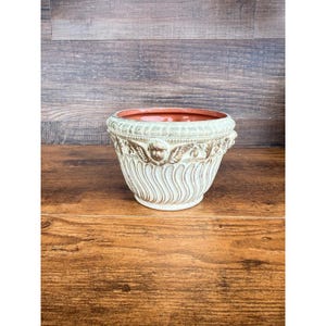 Puede incluir: Una maceta redonda de cerámica con un exterior de color crema que presenta un patrón texturizado de remolinos verticales y una banda decorativa de rostros de querubines esculpidos. El interior es liso y de color naranja terracota.