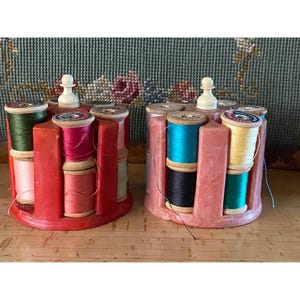 May include: Two circular rotating thread spool holders made of marbled plastic, one in red and one in pink. Each holder features a central white knob and contains multiple wooden spools of sewing thread in various colors including green, pink, blue, and yellow.