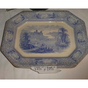 Puede incluir: Una fuente de servir octogonal antigua con un diseño de paisaje azul y blanco. La fuente mide 34,3 x 26,7 cm. La escena central representa un río, un puente y un castillo. El borde presenta motivos florales y arquitectónicos.