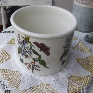 May include: A white ceramic planter with a floral design featuring purple and red flowers and green leaves. The planter has a wide, rounded shape and sits on a white lace doily. The design includes text in a script font.