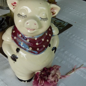 May include: A cream-colored ceramic pig figurine with closed eyes and long eyelashes. The pig wears a dark red scarf featuring white polka dots and blue accents. It has black painted hooves and a small dark spot on its side, with a dark pink flower.