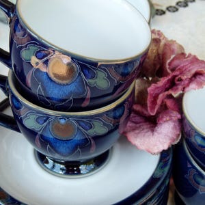 May include: A set of dark blue ceramic teacups and saucers featuring a white interior and a decorative floral pattern in shades of blue, green, and bronze. The cups have a curved handle and a pedestal base, with a matching floral design on the saucers.