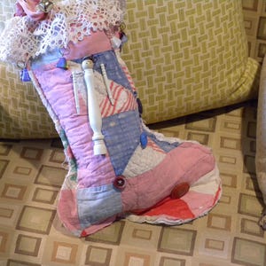 May include: A pink and blue patchwork stocking filled with various items, including a wooden doll, buttons, and lace. The stocking is sitting on a patterned couch.