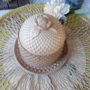 May include: A beige, textured ceramic jar with a lid featuring a rose-shaped handle, resting on a matching saucer. The jar and saucer are set on a yellow tray with a decorative net overlay, with a white flower and other decorative elements in the background.