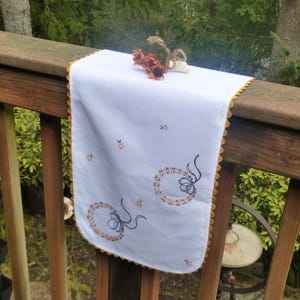 May include: A white table runner with embroidered wreath designs and gold scalloped edges. The runner is decorated with small star-shaped embroidery and is draped over a wooden railing. A small floral arrangement sits on top.