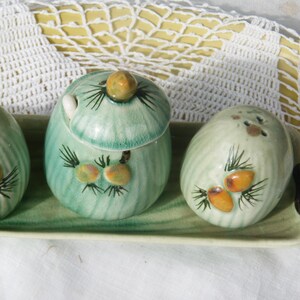 Puede incluir: Un juego de salero y pimentero de cerámica vintage y un recipiente con tapa sobre una bandeja rectangular. El juego está pintado en tonos verdes con detalles marrones y amarillos. El salero tiene dos agujeros.