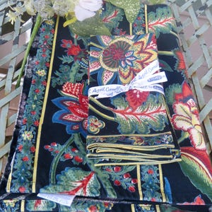 May include: A set of floral patterned table linens, including a tablecloth and folded napkins. The design features large red, blue, and yellow flowers on a black background. The napkins are tied with a white ribbon that reads "April Cornell".
