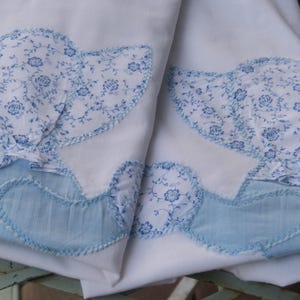 Könnte beinhalten: Weißer Stoff mit blau floral gemusterten Applikationen. Die Applikationen haben die Form von Hauben und sind mit einer blau genähten Bordüre versehen. Die Haubenapplikationen haben eine hellblaue Stoffbasis.