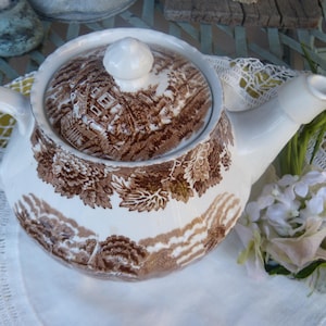 May include: White ceramic teapot with a brown landscape pattern on the body and lid. The teapot has a curved handle and spout. The lid has a small knob. The teapot is decorated with floral and landscape designs.
