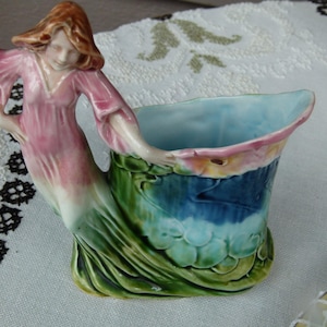 May include: A ceramic figurine vase featuring a sculpted figure with auburn hair wearing a flowing pink dress that transitions into a green base. The figure holds the edge of a curved, shell-like vessel with a light blue interior and green exterior.