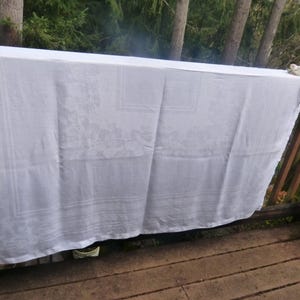 May include: A white tablecloth with a floral pattern is draped over a wooden railing. The tablecloth is made of a textured fabric and hangs down, revealing the intricate design. The background features trees and a wooden deck.