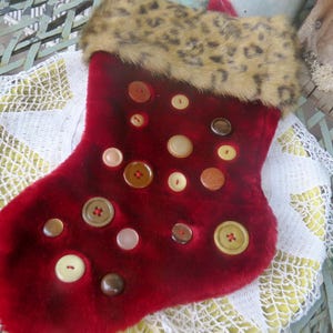 May include: A red Christmas stocking with a faux leopard fur cuff and various buttons. The stocking is decorated with an assortment of buttons in shades of brown, cream, and tan. The stocking is likely handmade and adds a festive touch to holiday decor.