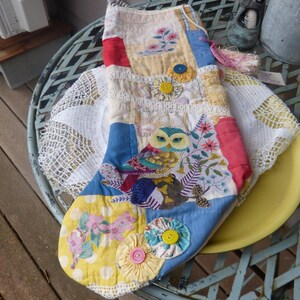 May include: A handmade patchwork oven mitt featuring an owl design. The mitt is made of various fabric scraps in blue, yellow, red, and floral patterns, adorned with decorative buttons and lace trim. A tassel hangs from the top.
