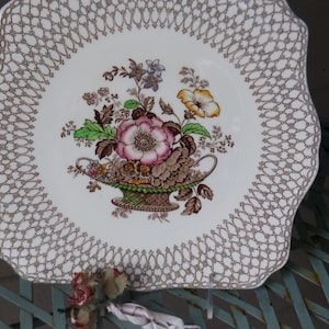 Puede incluir: Un plato de servir de cerámica blanca con un patrón de encaje marrón alrededor del borde. El centro presenta un diseño floral con flores rosas, amarillas y verdes en un jarrón marrón y dorado.