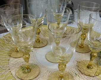 Pode incluir: Conjunto de seis copos de vinho de vidro transparente com hastes e bases de cor âmbar. Os copos estão dispostos sobre um crochê branco em uma bandeja amarela.
