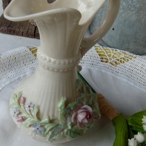 May include: A cream-colored ceramic pitcher featuring a fluted neck, a beaded band, and a handle. The base is decorated with three-dimensional floral accents, including a prominent pink rose, small blue flowers, and green leaves.