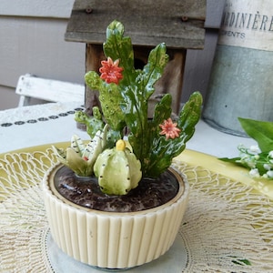 May include: A ceramic planter featuring a glossy, ribbed cream-colored base filled with dark brown soil. It contains a green cactus with tall, wavy pads, two small orange flowers, a round light green cactus, and a small white succulent with stripes.