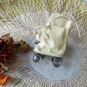 May include: A miniature cream-colored roller skate featuring a smooth synthetic upper, white laces, and four small silver-toned metal wheels. The skate is constructed with a high-top design and includes a visible metal chassis connecting the wheels.
