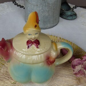 Puede incluir: Una tetera de cerámica con forma de payaso, con tapa en forma de sombrero amarillo, pajarita roja y pantalones azules. El pico tiene forma de figura roja parecida a un cerdo y el asa es curva. La tetera presenta un acabado brillante.
