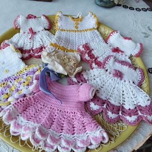 May include: A collection of small, handmade crochet dress ornaments in various colors including white, pink, yellow, and purple. Each miniature dress features intricate stitching, scalloped hems, and decorative accents like small buttons or ribbons.