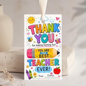 May include: A rectangular gift tag with a white lined paper background featuring colorful illustrations of a sun, bee, butterfly, apple, and crayons. The text reads: THANK YOU for making learning fun! YOU ARE THE BEST TEACHER EVER! From: Emma.