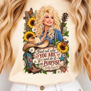 May include: A cream-colored t-shirt featuring a graphic of a blonde woman, a cowboy hat, a guitar, sunflowers, butterflies, and a cowboy boot. The text reads: Find out who YOU ARE and do it on PURPOSE - Dolly Parton, with heart accents.
