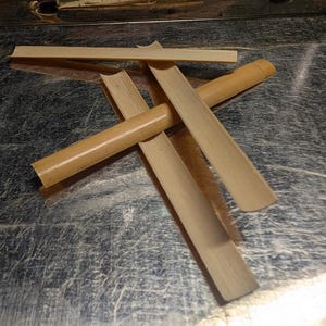 May include: A collection of light brown, reed-like objects, possibly reeds or parts of a musical instrument, are arranged on a metallic surface. The objects are of varying lengths and shapes, with some appearing cylindrical and others flat.