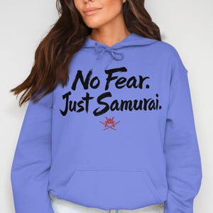 Könnte beinhalten: Ein periwinkelfarbener Hoodie mit Kapuze und Kängurutasche. Auf der Vorderseite steht in schwarzer Pinselbeschriftung der Text "No Fear. Just Samurai." mit einer roten Grafik mit gekreuzten Schwertern darunter.