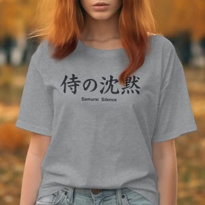 May include: A grey t-shirt featuring the Japanese phrase "Samurai Silence" in black lettering. The text is centrally positioned. The shirt is made of a soft fabric.