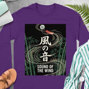 May include: A long-sleeved purple shirt featuring a graphic with Japanese text and the English phrase "SOUND OF THE WIND." The graphic depicts bamboo stalks, flowing lines, and a wind chime. The shirt is made of a soft material.