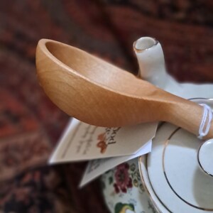 May include: A wooden spoon rests on a floral-decorated teapot. The spoon is light brown and has a curved bowl. The teapot is white with gold trim and a colorful floral pattern. A tag is partially visible.