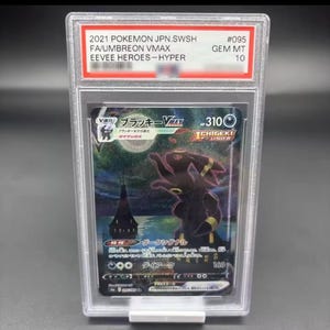 Puede incluir: Una carta Pokémon Umbreon VMAX del set Eevee Heroes, calificada y protegida en un estuche transparente. La carta está calificada como GEM MT 10. La ilustración muestra a Umbreon en una escena oscura a la luz de la luna. El texto es japonés.