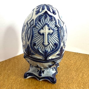 May include: A decorative ceramic egg featuring a raised white cross centred on a blue and white patterned background. The egg is supported by a matching blue and white pedestal base with ornate, scalloped edges and intricate relief detailing.