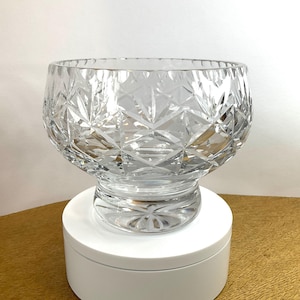 May include: A clear, cut-glass bowl with a wide, rounded shape. The bowl features a detailed diamond and starburst pattern, sitting on a short, pedestal base. The glass reflects light, creating a sparkling effect. The bowl is displayed on a white surface.