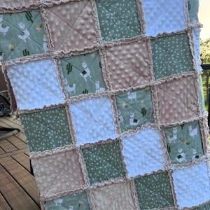 May include: A handmade patchwork quilt featuring squares of white, sage green, and tan fabrics. Some squares have a dotted texture, while others display llama and cactus patterns. The quilt has a ruffled edge, adding a soft, textured look.