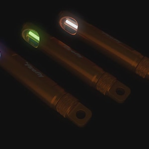 Może przedstawiać: Trzy cylindryczne metalowe znaczniki Firefly z teksturowanymi sekcjami chwytnymi i pętlami mocującymi na jednym końcu. Każdy znacznik posiada świecącą wkładkę u góry, emitującą odpowiednio niebieskie, zielone i białe światło.
