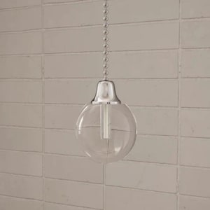 May include: A clear glass globe pendant light fixture featuring a polished chrome metal cap and a matching silver-toned ball chain pull. The spherical glass shade is transparent, revealing a central cylindrical light socket component inside.