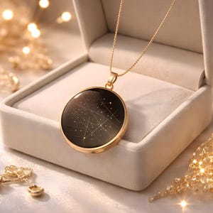 Könnte beinhalten: Eine goldfarbene Halskette mit einem runden Anhänger, der ein Sternbilddesign auf dunklem Hintergrund zeigt. Der Anhänger befindet sich in einer weißen Schmuckschatulle. Die Halskette ist ein zartes Schmuckstück.