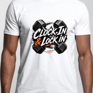 Może przedstawiać: Biały t-shirt z krótkim rękawem z grafiką przedstawiającą dwa czarne hantle skrzyżowane za napisem CLOCK IN & LOCK IN w kolorze białym i pomarańczowym. Poniżej głównego tekstu znajduje się mniejszy pomarańczowy napis FOCUS. DISCIPLINE. RESULTS. NO EXCUSES.