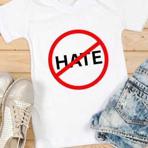 Puede incluir: Una camiseta blanca de manga corta con un gráfico de un círculo rojo con una línea diagonal, superpuesto a la palabra HATE en letras mayúsculas negras en negrita. La camiseta se muestra junto a un par de zapatillas plateadas y unos vaqueros azules.