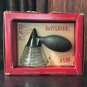 May include: A red and white box with a clear glass bottle with a black rubber bulb attached. The box says "Devilbiss" and "For Applying Any Solution". The bottle is labeled "$1.00".
