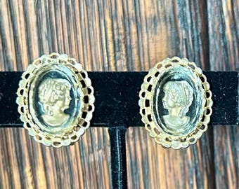 Könnte beinhalten: Ein Paar Vintage-Ohrringe mit Kamee. Jeder Ohrring zeigt das Profil einer Frau in cremefarbenem Relief vor dunklem Hintergrund, eingebettet in einen ovalen, goldfarbenen Rahmen mit dekorativem Rand. Die Ohrringe werden auf einem schwarzen Ständer vor Holzmaserung präsentiert.