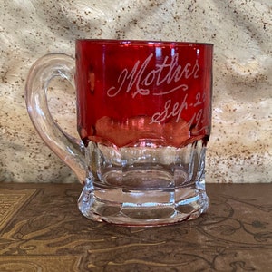May include: A small, red glass mug with a handle. The mug has the words "Mother Sep. 2" etched into the side.